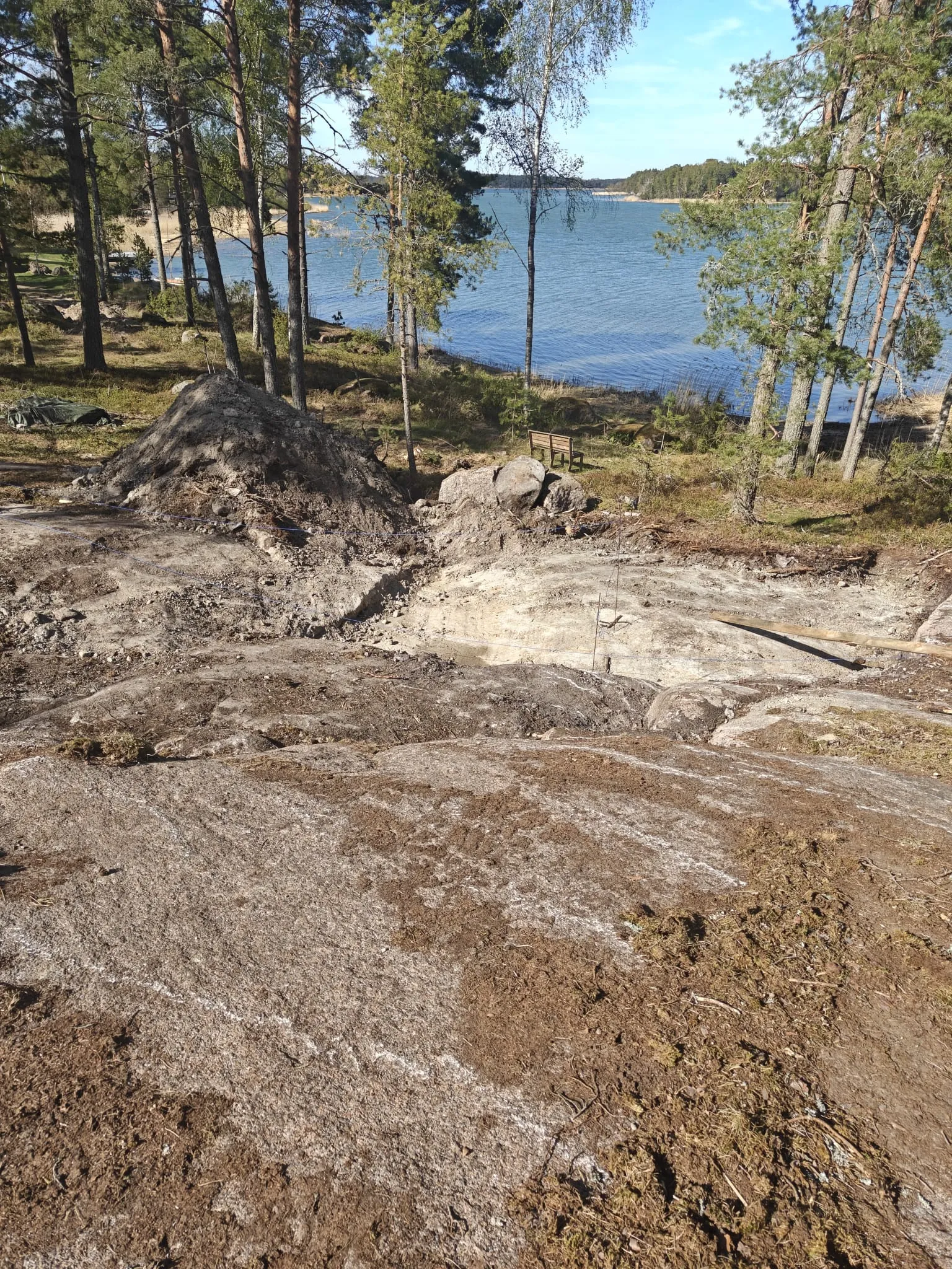 Rakennusalan tontti järven rannalla, jossa on kaivettua maata, kalliota ja puita.