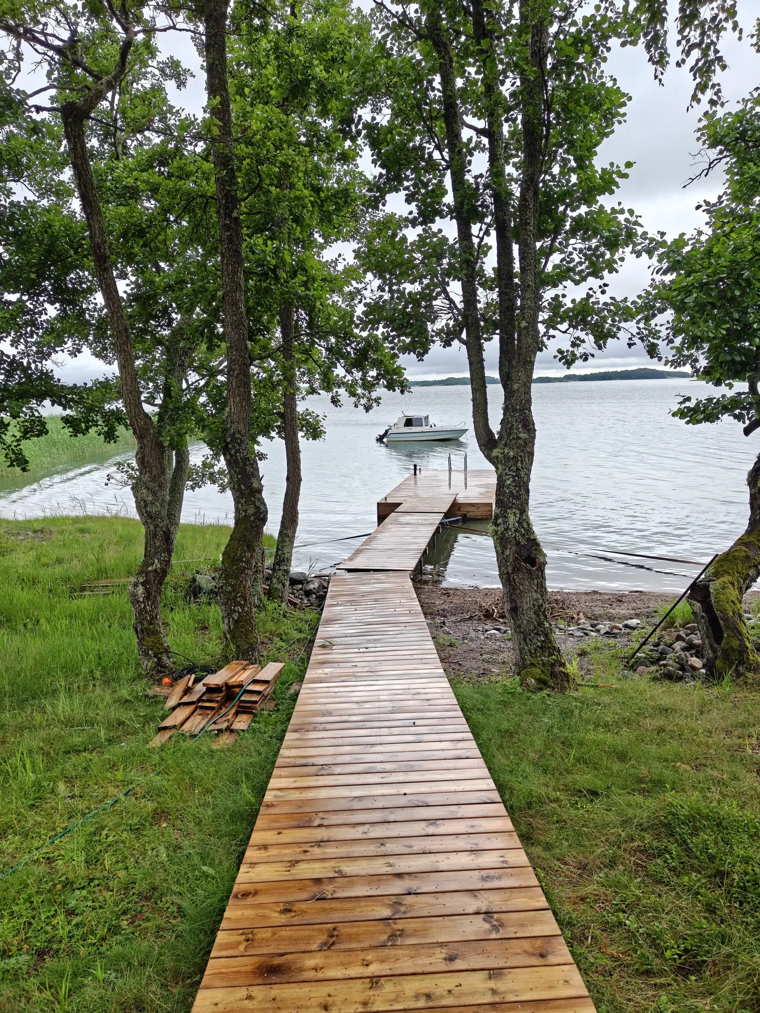 Puinen laituri johtaa järvelle, jossa on vene, ja laiturin vieressä on rakennusmateriaaleja.