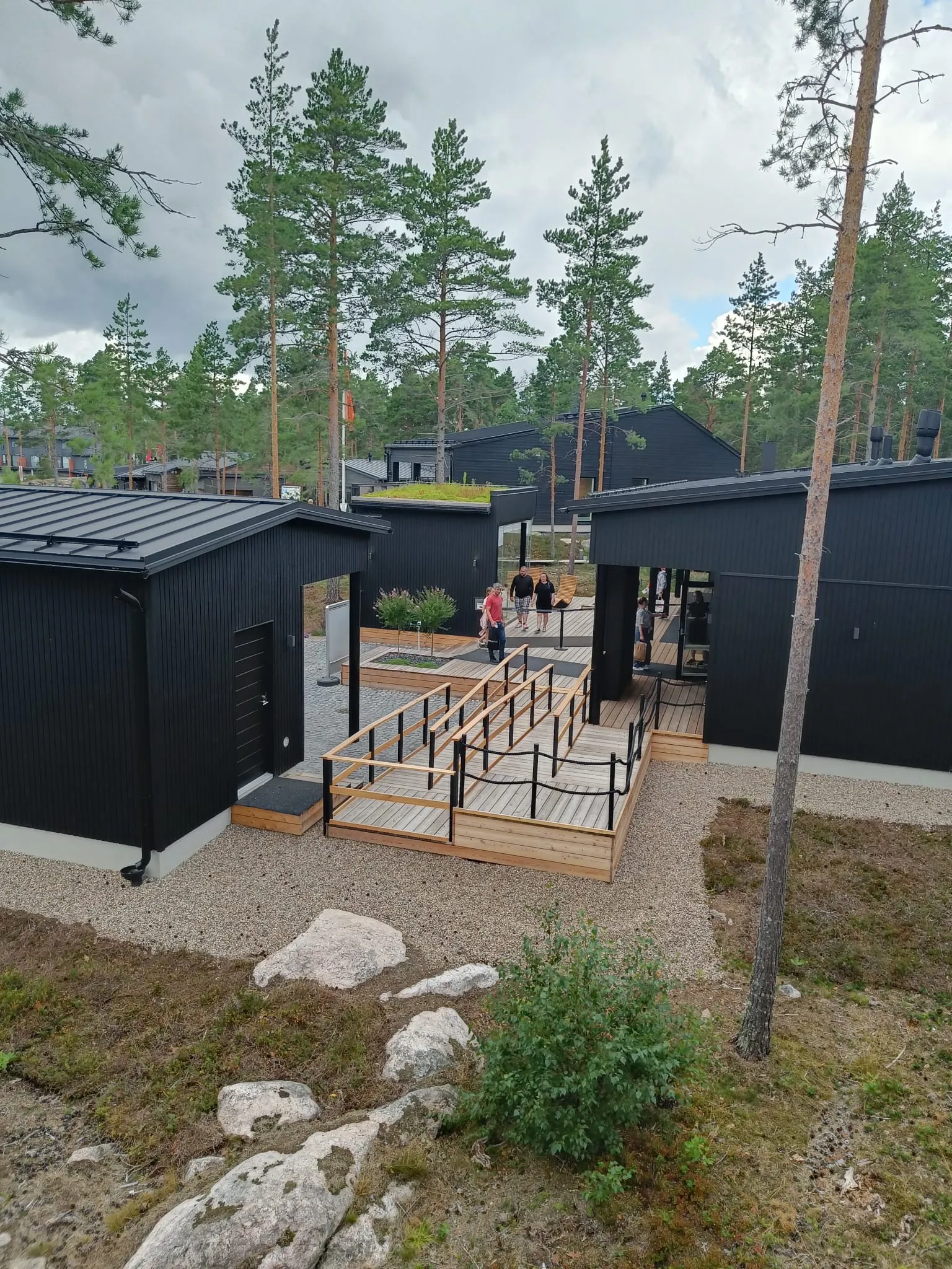 Mustia moderneja rakennuksia, joissa on puinen luiska ja sorapolut, ympäröi mäntymetsä, ja ihmisiä kävelee rakennuskohteiden välissä.