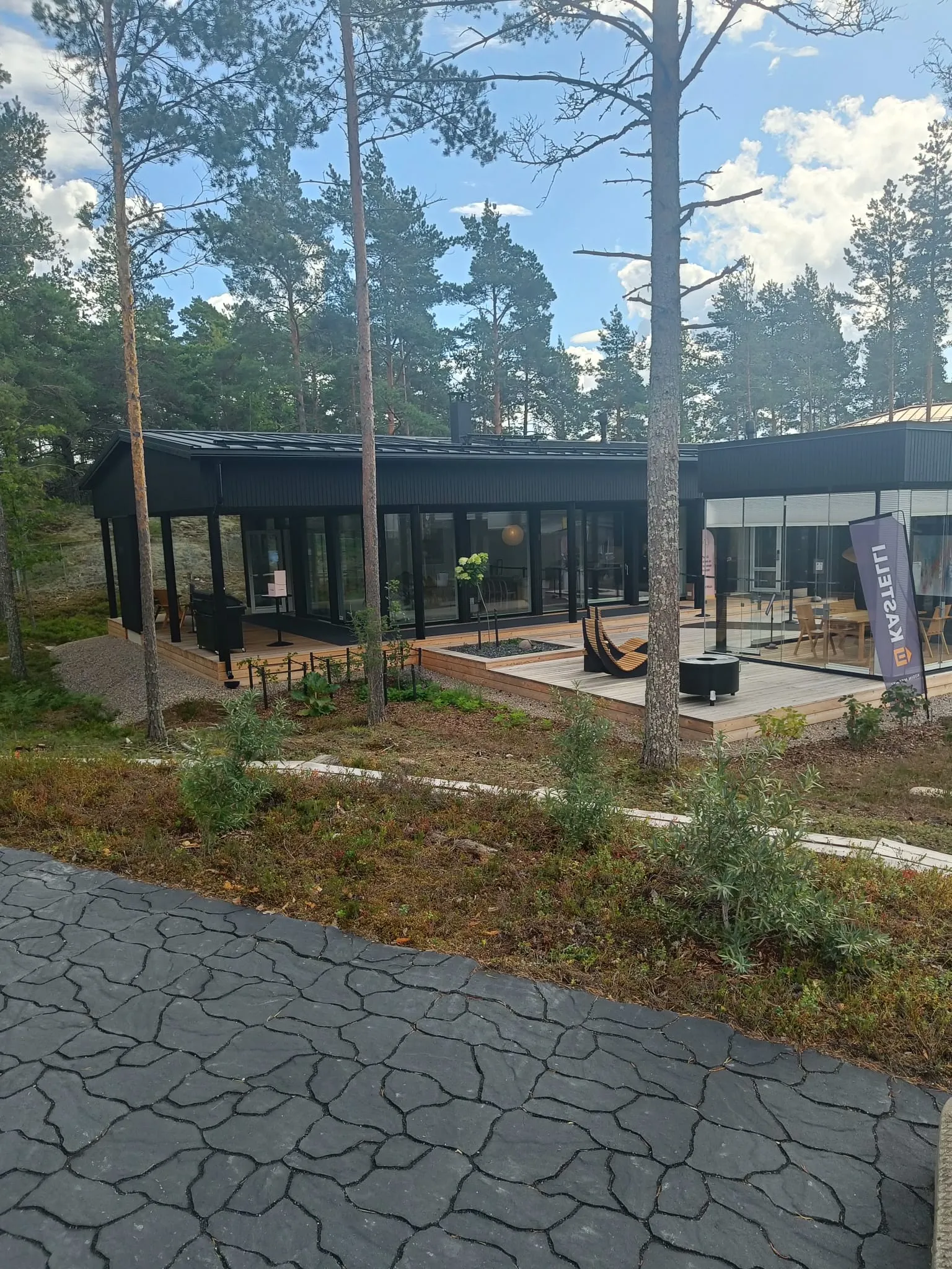 Nykyaikainen musta talo suurilla ikkunoilla ja puuterassilla metsäisessä ympäristössä, joka on esimerkki rakentamisesta.