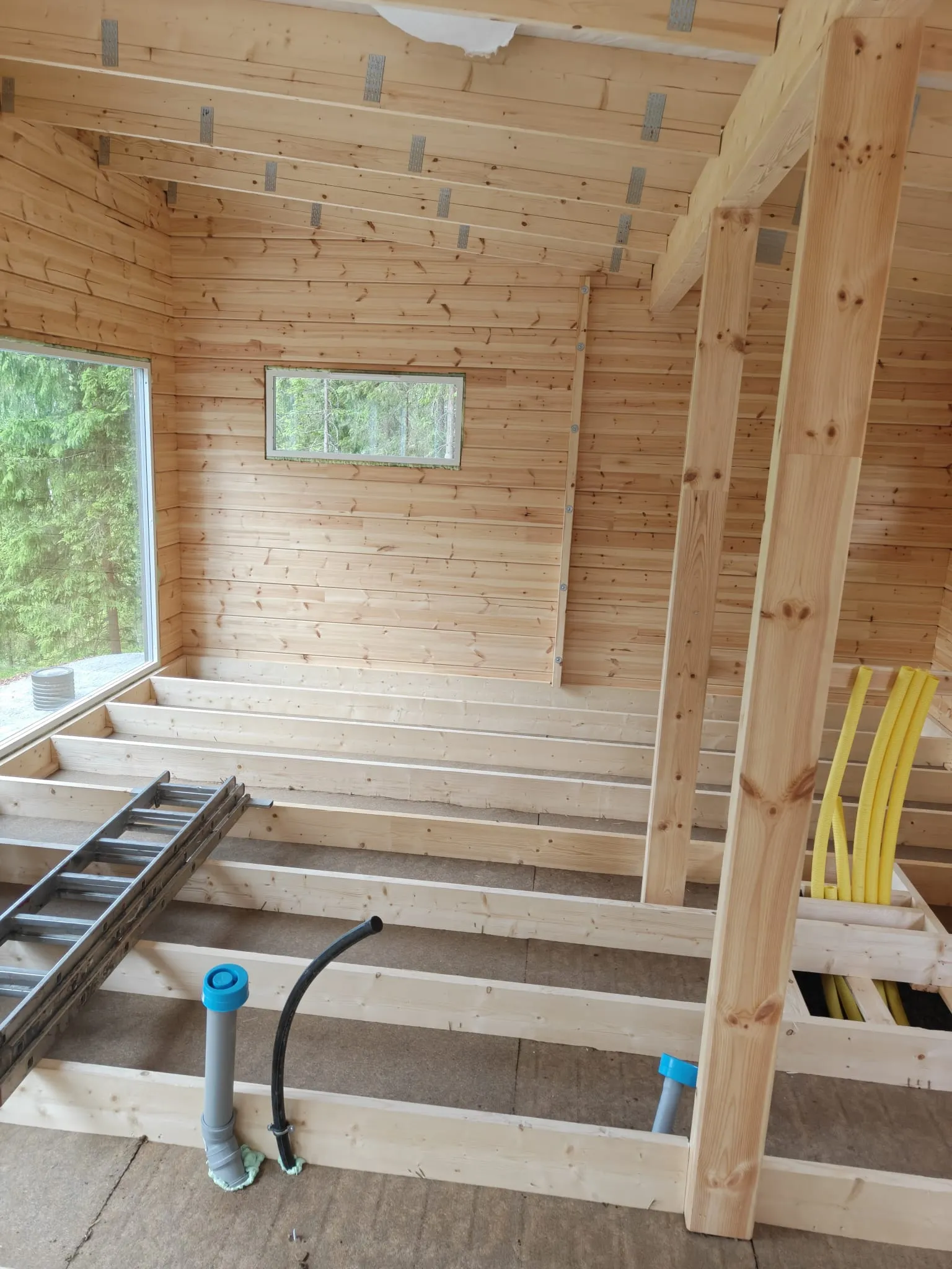 Keskeneräinen puurakennus, jossa näkyy lattiavasat, kattopalkit ja ikkunat.