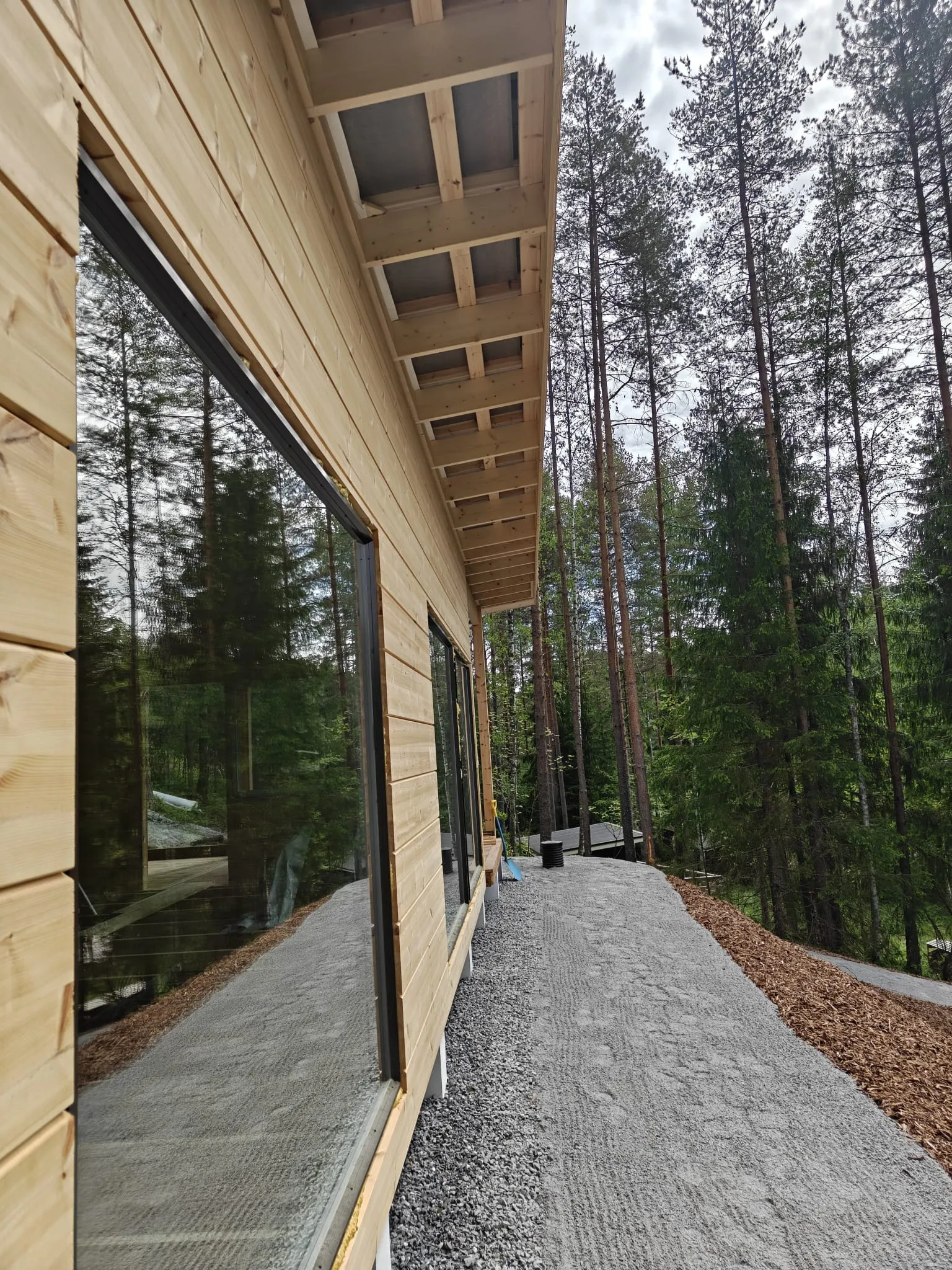 Rakentamisen alla oleva puinen rakennus, jonka suuret ikkunat heijastavat metsää, seisoo sorapolun vieressä.