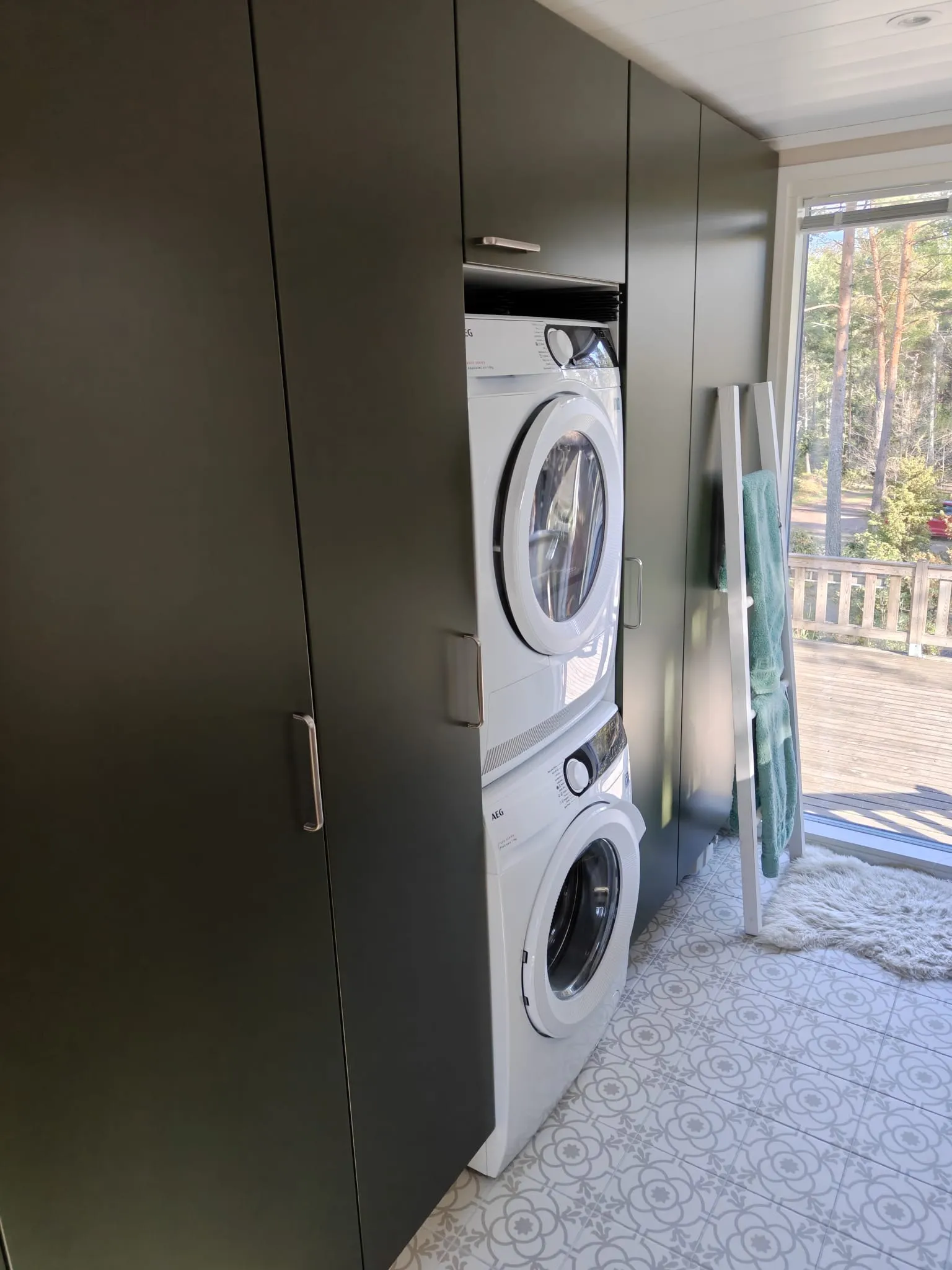 Pyykinpesuhuoneessa on tummanvihreitä kaappeja, pinottu pesukone ja kuivausrumpu, sekä ikkuna, josta avautuu näkymä terassille ja metsään, mikä on osa huoneen rakentamista.