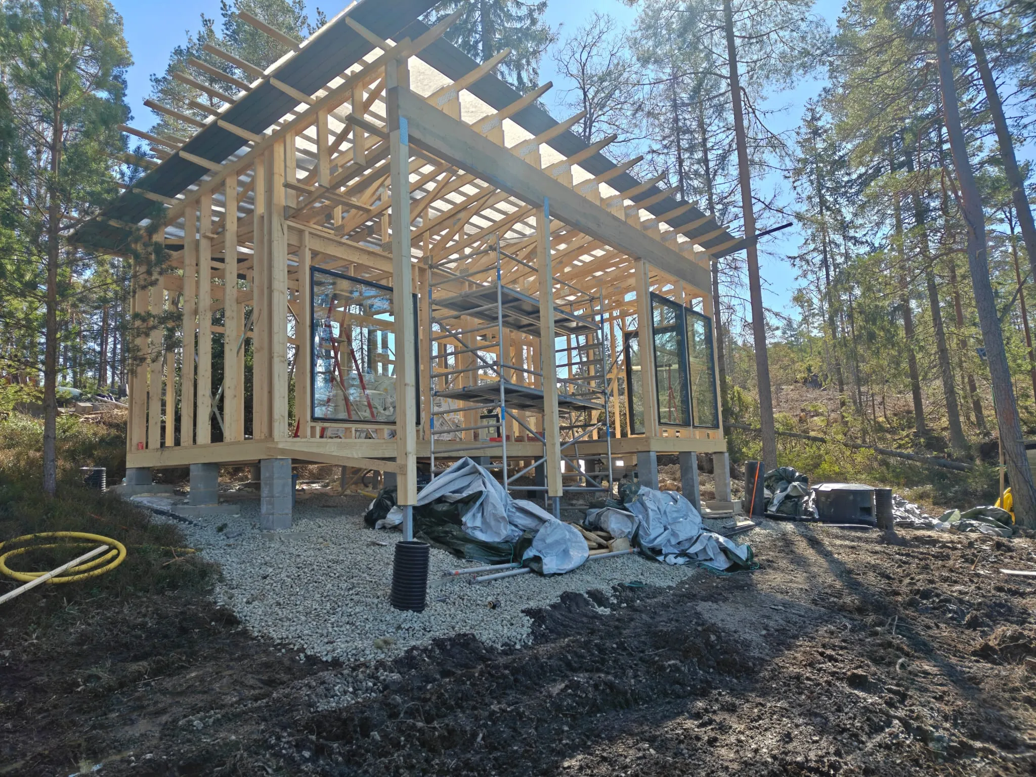 Metsäisessä ympäristössä on puurunkoinen talo rakentamisen alla, sen kattorakenne on osittain valmis ja ympärillä on rakennustyömaan materiaaleja.