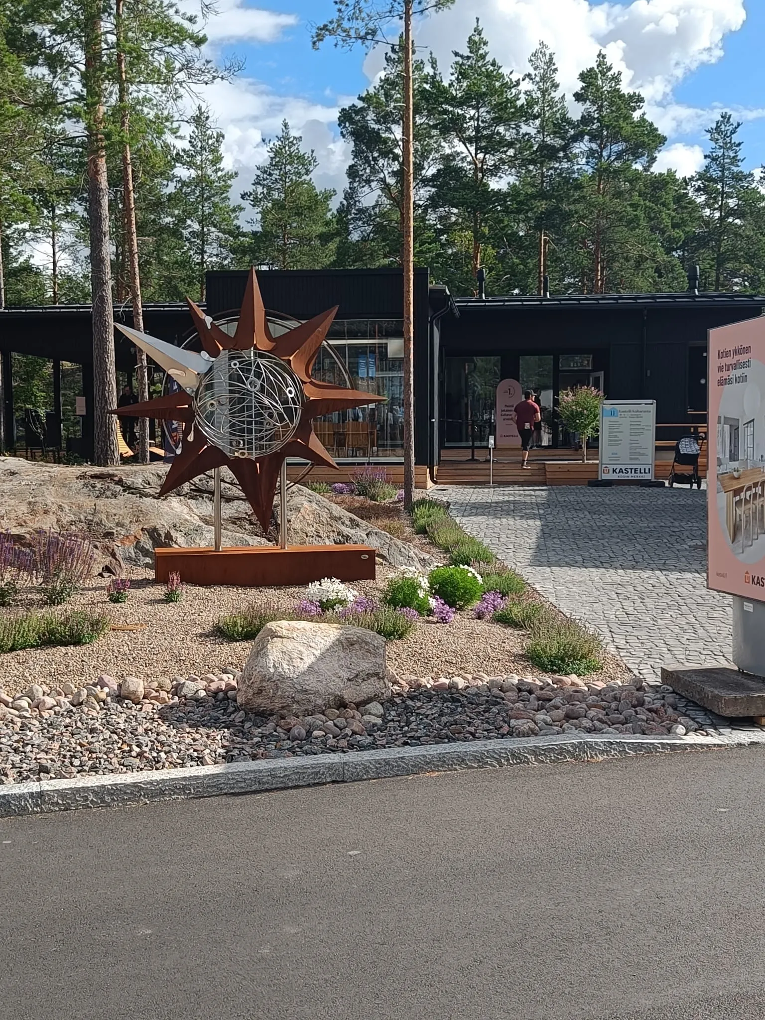 Moderni rakennus ja sen edustalla oleva maisemoitu piha-alue, jossa on suuri metalliveistos, kuvastavat rakennusalan suunnittelua ja rakentamista.