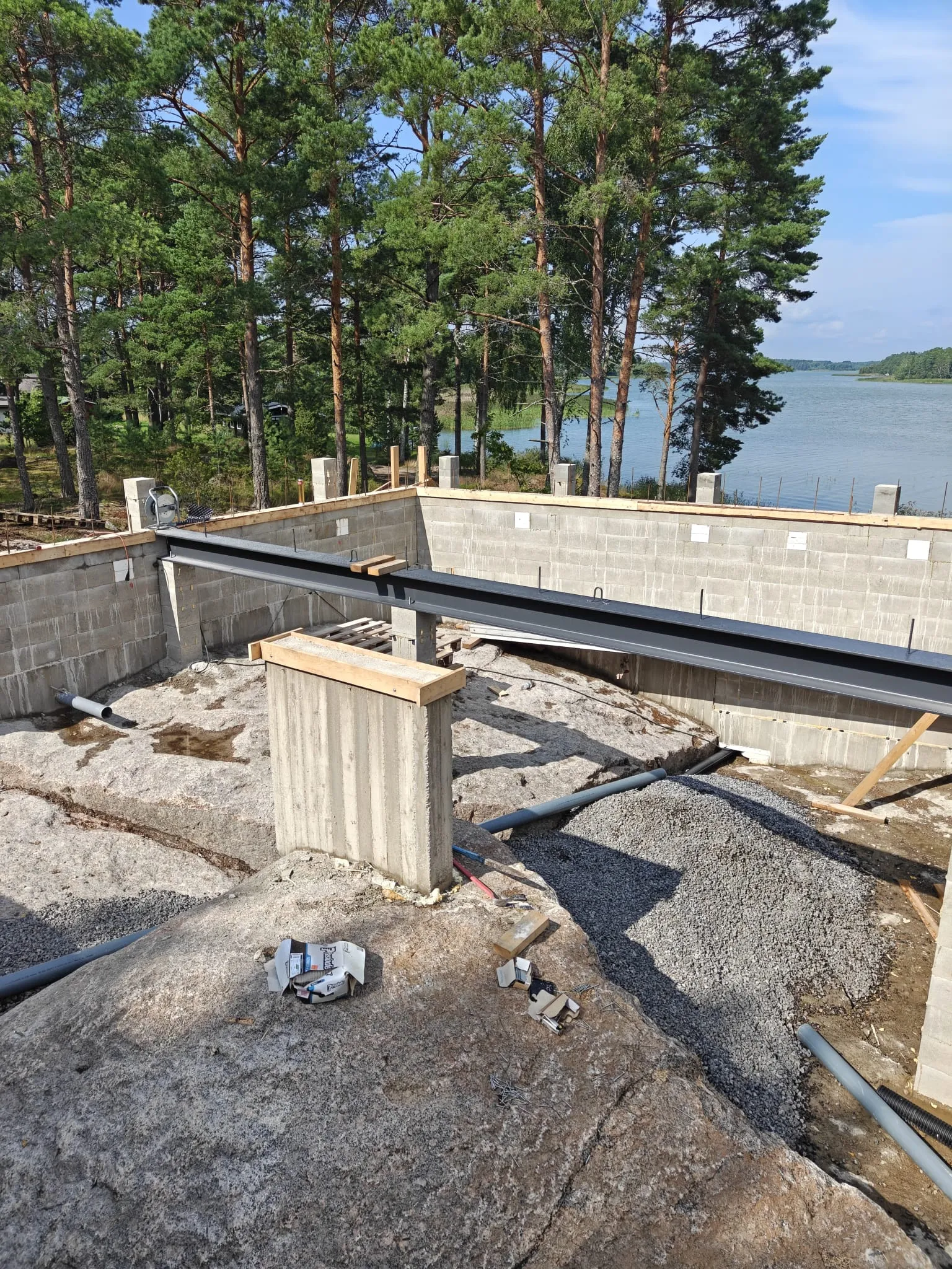 Rakennustyömaa, jossa on talon perustukset, betoniharkkoseinät ja teräspalkki, taustalla metsäinen järvimaisema.