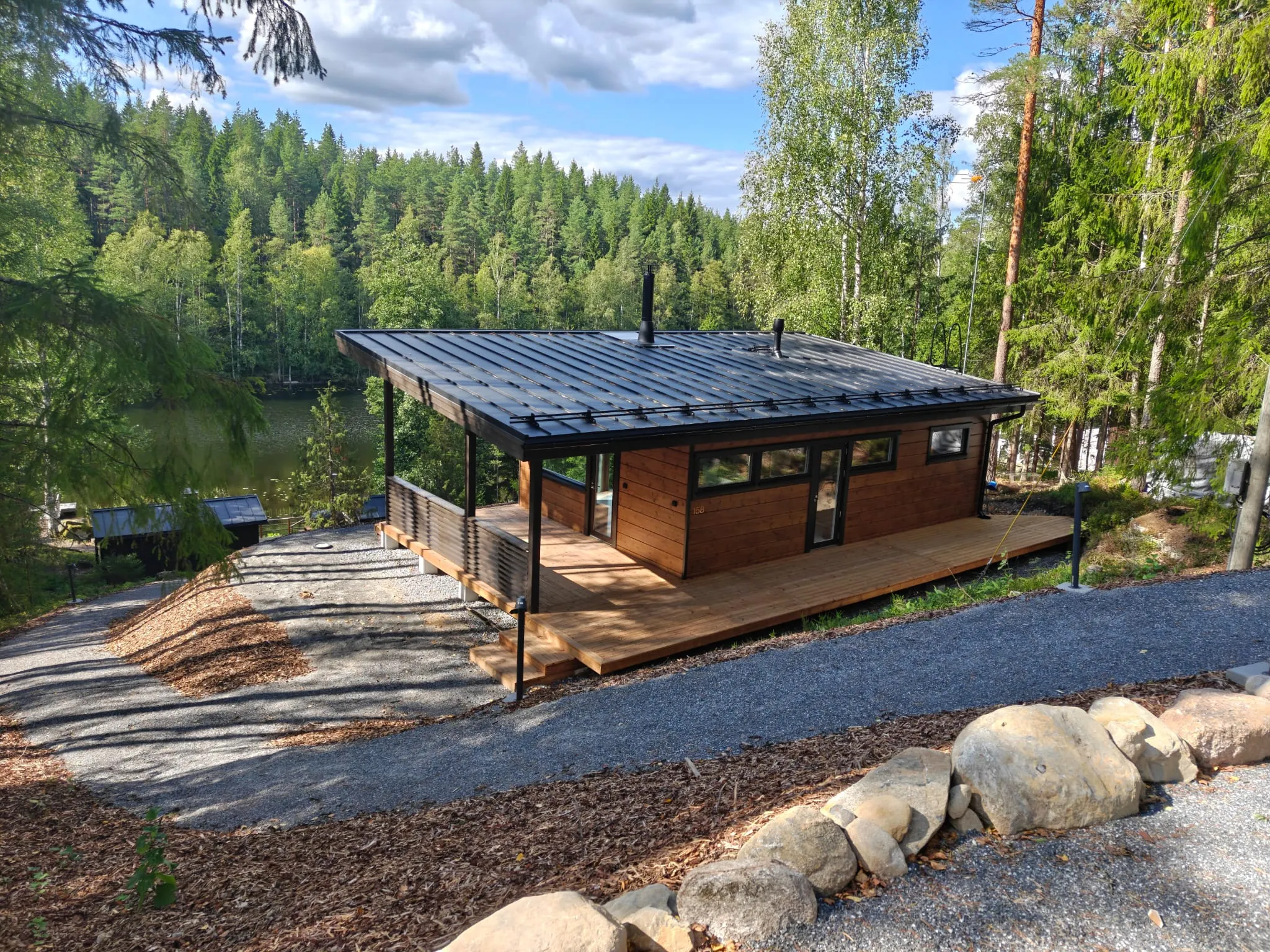 Metsäisellä rannalla sijaitseva moderni puurakennus, jossa on tumma peltikatto ja tilava puuterassi.