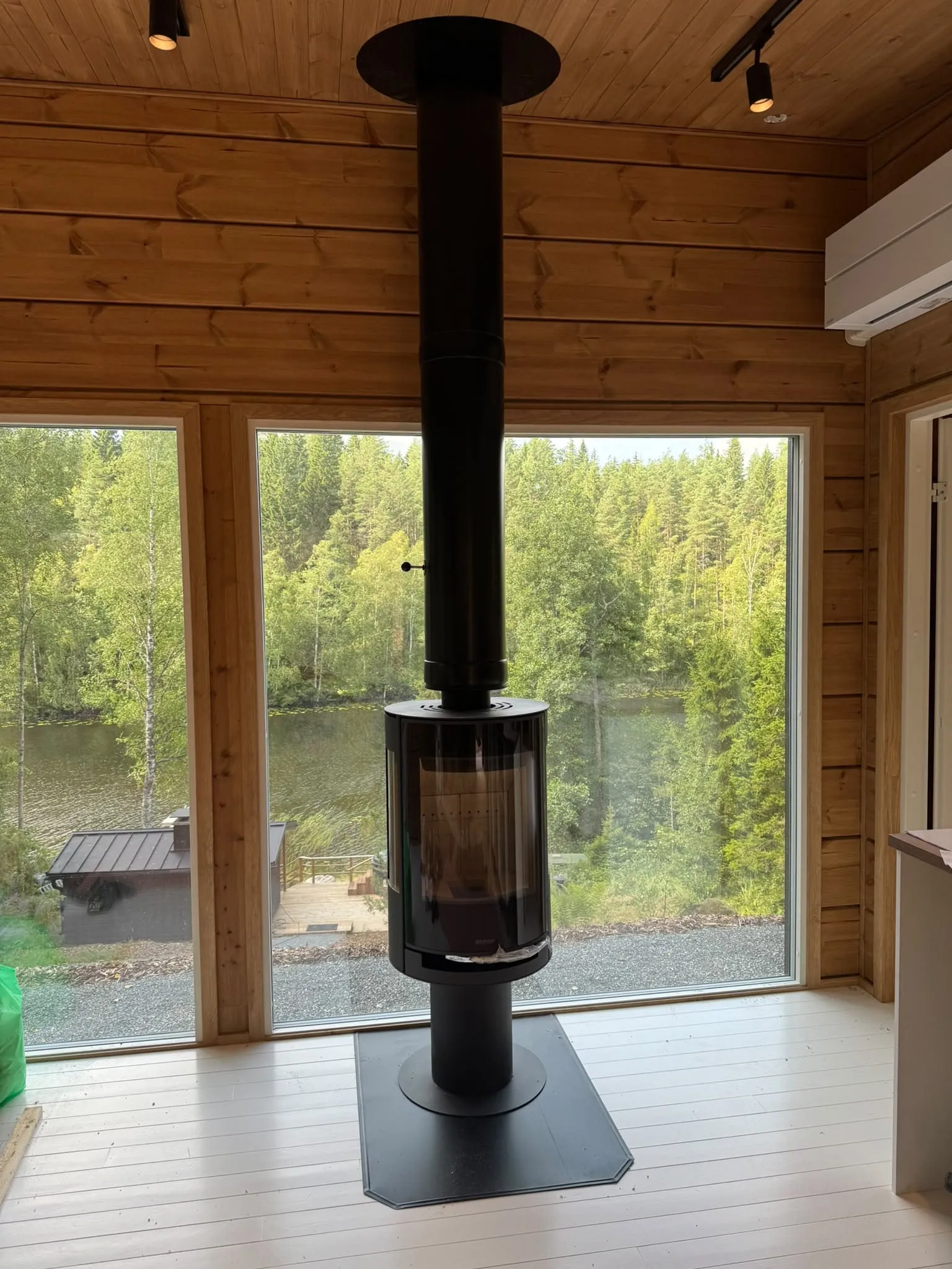 Moderni musta takka ja savupiippu puuseinäisessä rakennuksessa, josta avautuu näkymä järvelle ja metsään.