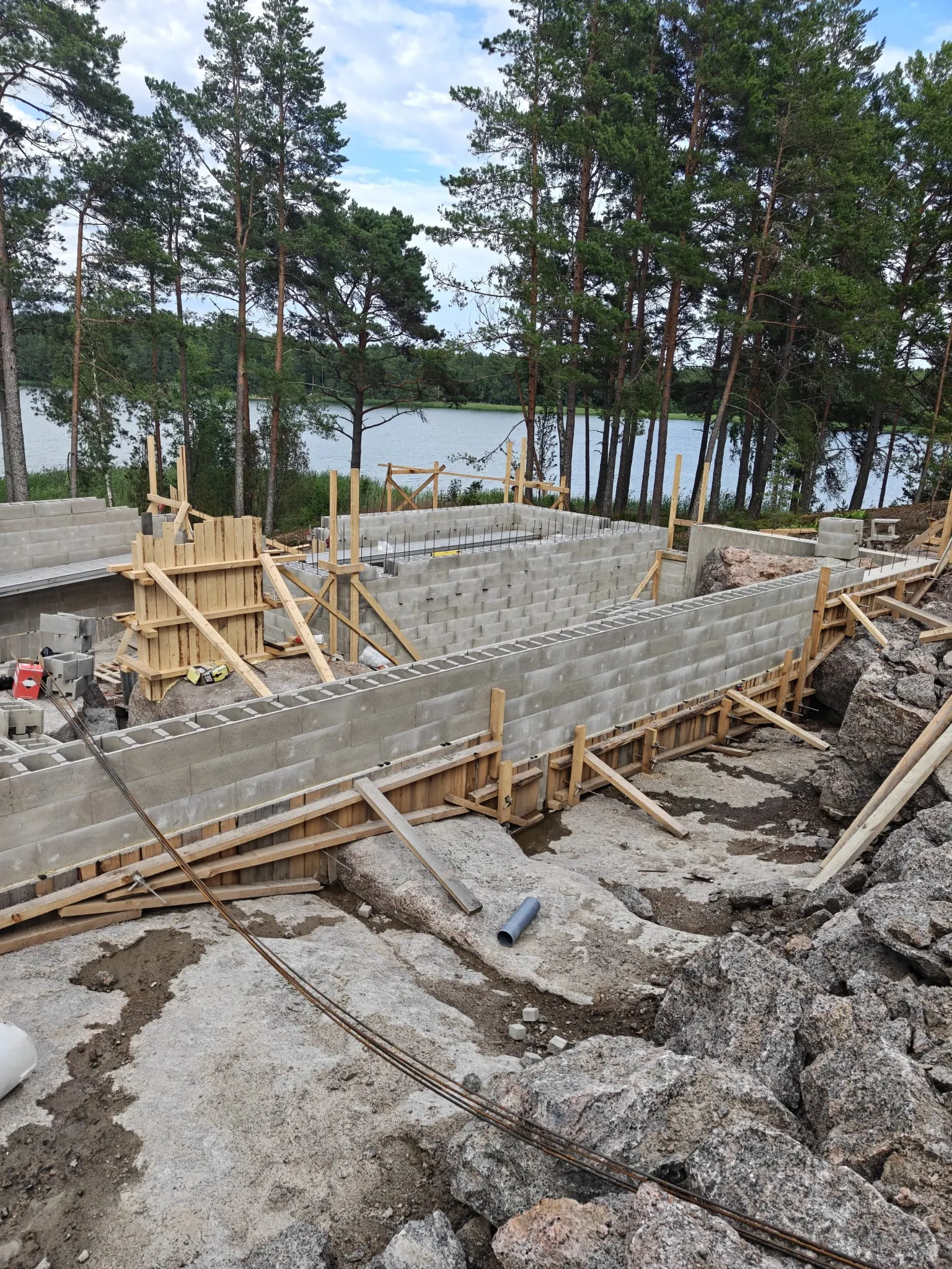 Rakennustyömaa, jossa rakennetaan talon perustusta betoniharkoista ja puumuoteista järven rannalla.